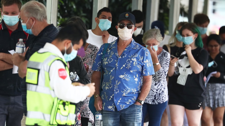 A queue of people wearing masks stand before a man in a high vis vest holding a clipboard