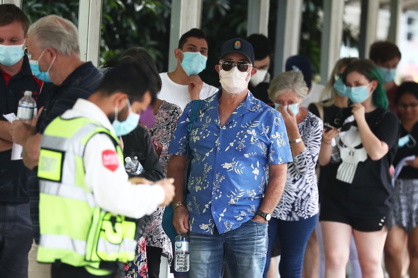 A queue of people wearing masks stand before a man in a high vis vest holding a clipboard