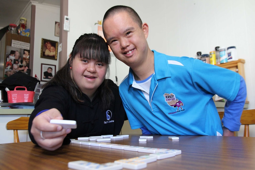 Jess and Jordy sit close together smiling, Jess' arm around Jordy and she is holding a domino piece above their domino game