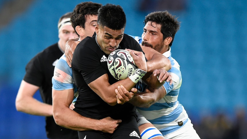 An All Blacks player holds the ball to his chest as he is tackled by two Argentine opponents.