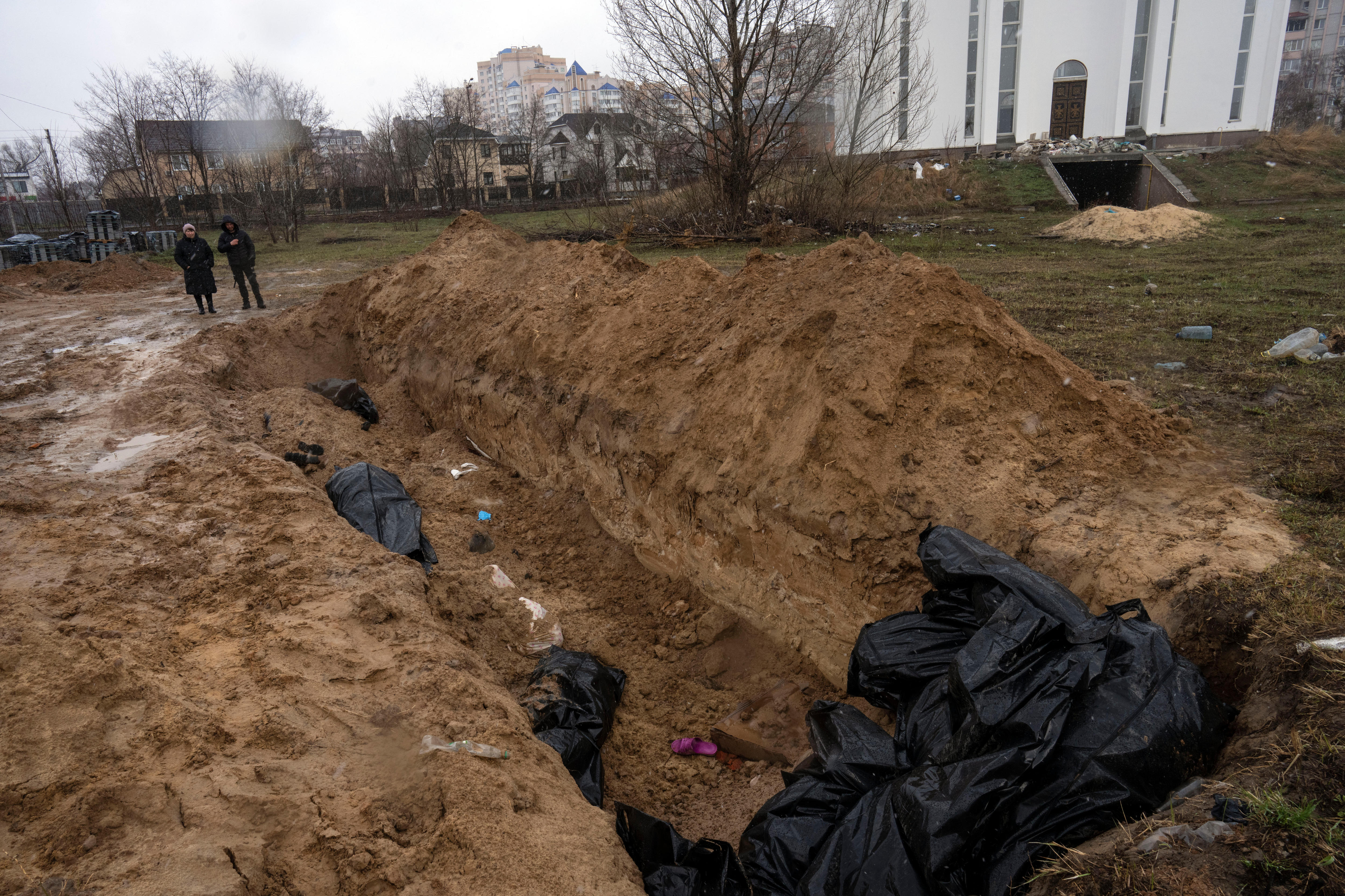 Two people dressed in black stand next to a mass grave.