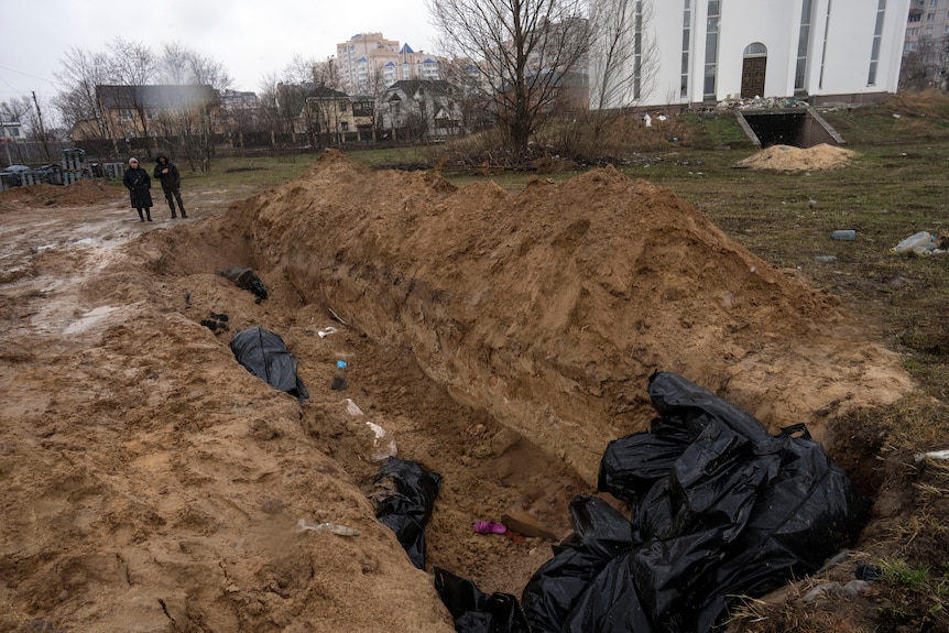 Two people dressed in black stand next to a mass grave.