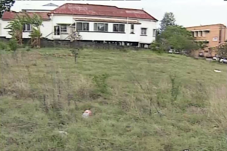 A vacant lot with overgrown grass and some rubbish in front of a timber house and another brick building.