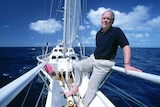 A white man stands on a sailboat while looking out to sea.