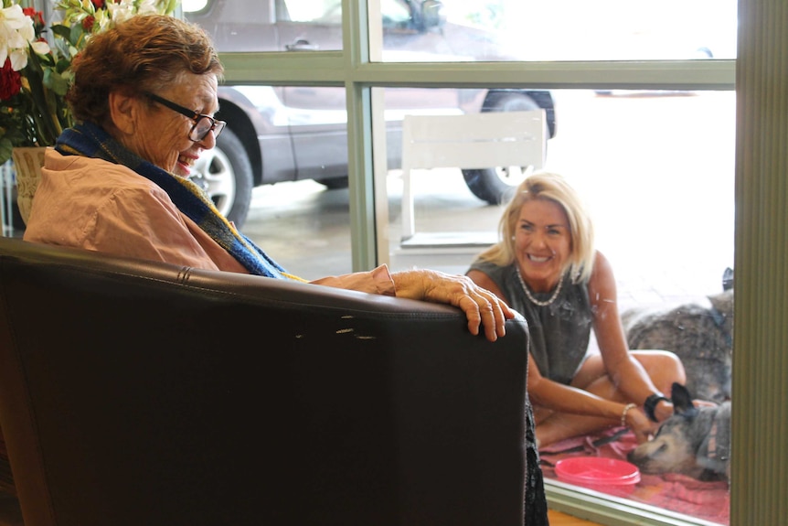 Older woman sits in armchair in front of window with a younger woman behind it, smiling at each other