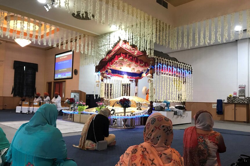 Women in colourful clothing and long scarves sit in front of a colourfully decorated canopy, white flowers hang from the ceiling