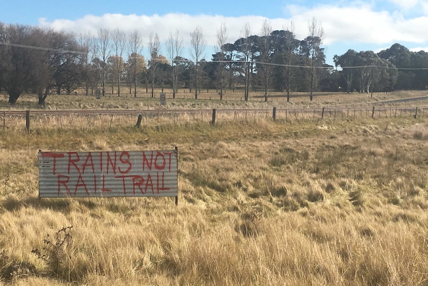 A corrugated iron sign  in a paddock near a rail track has trains not rail trail painted in red.