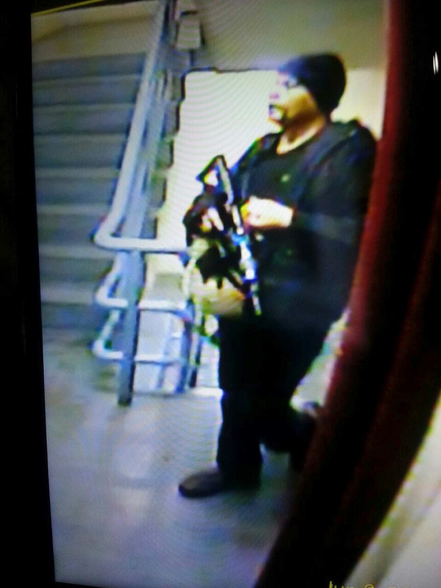 A man wearing black clothes and a beanie holds a large gun in a stairway.