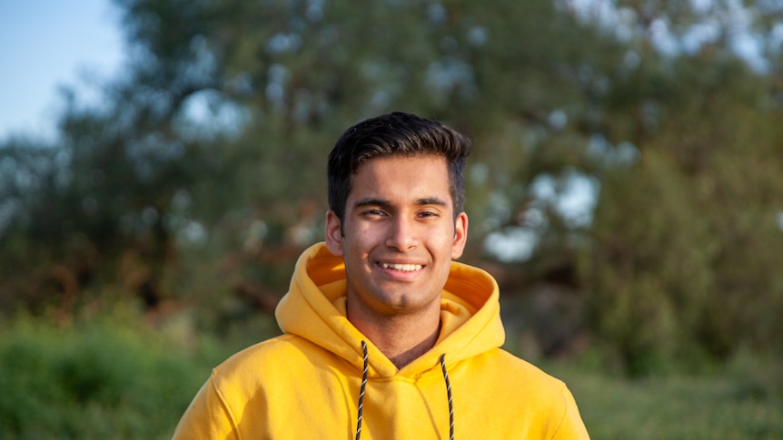 A young man wearing a yellow hoodie, smiling in a park