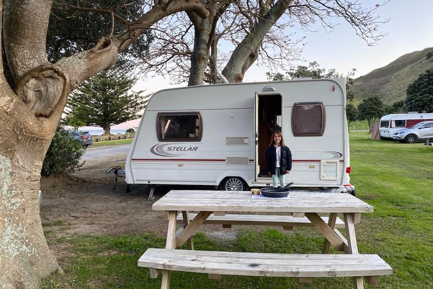 Her daughter stands in the door of the caravan, parked on grass by a bay, with a picnic table in front