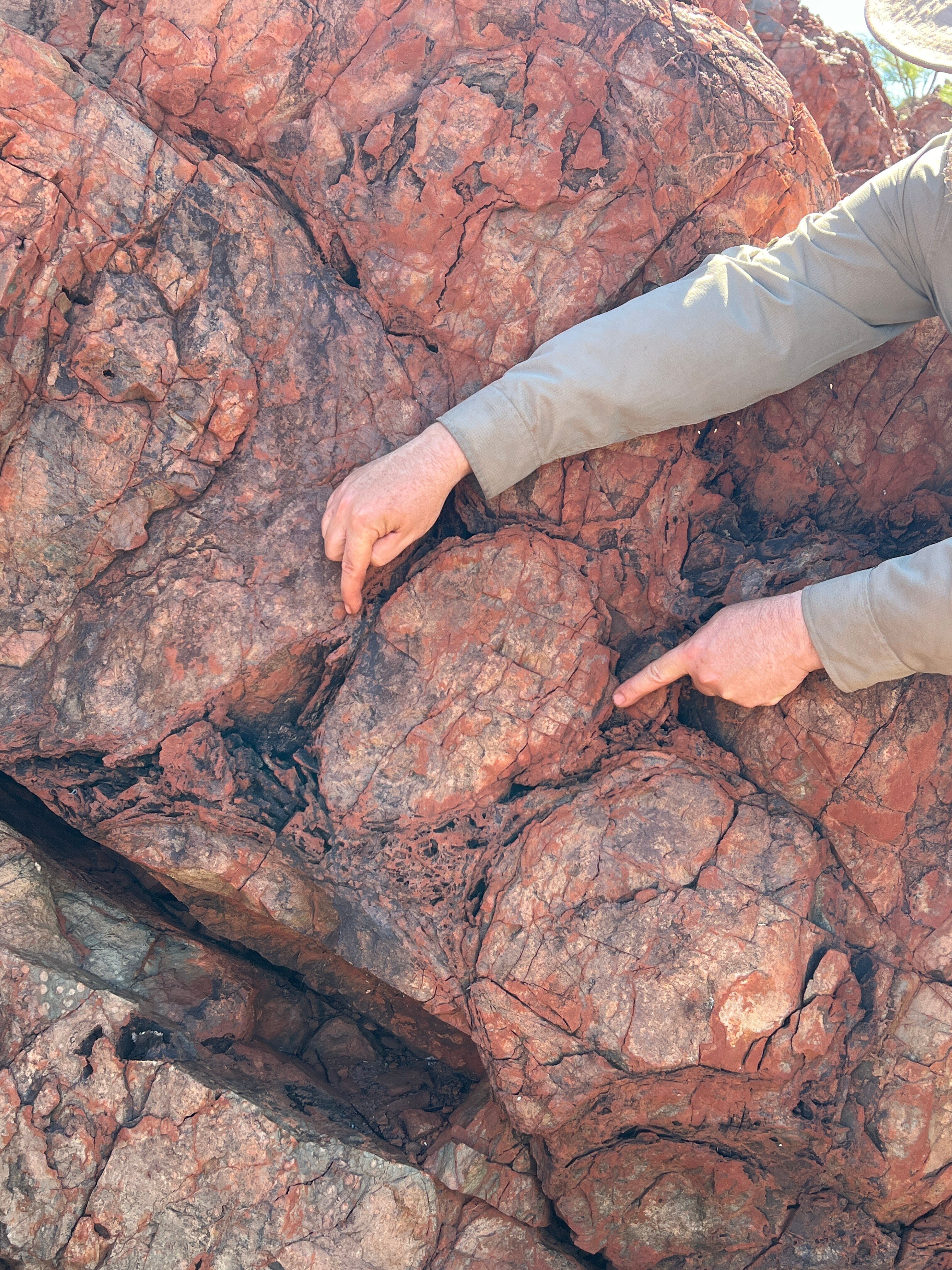 Two hands point to a circular formation on reddish rock.