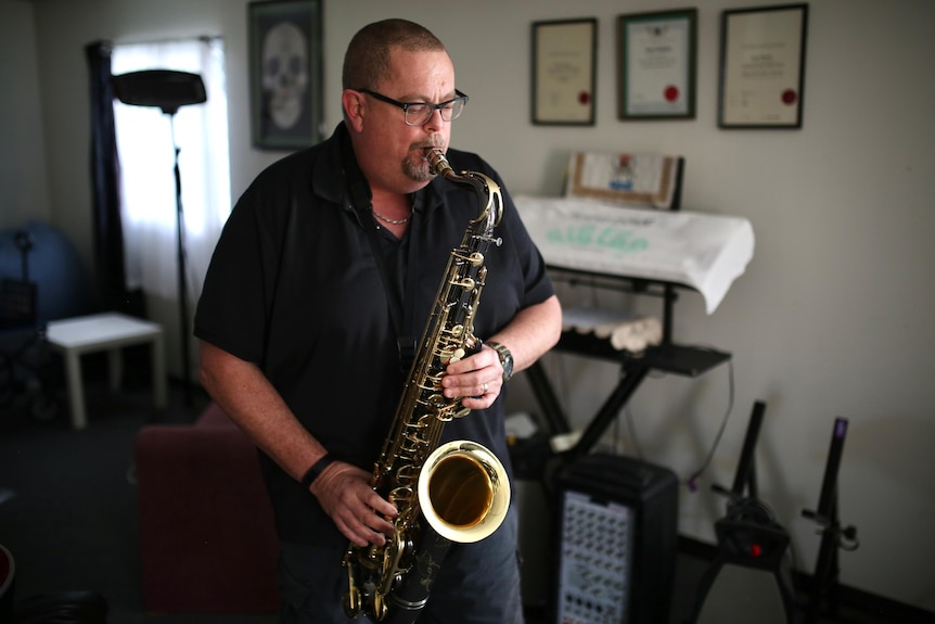 Andrew wears a black tshirt playing a saxophone in a music room in a suburban house.