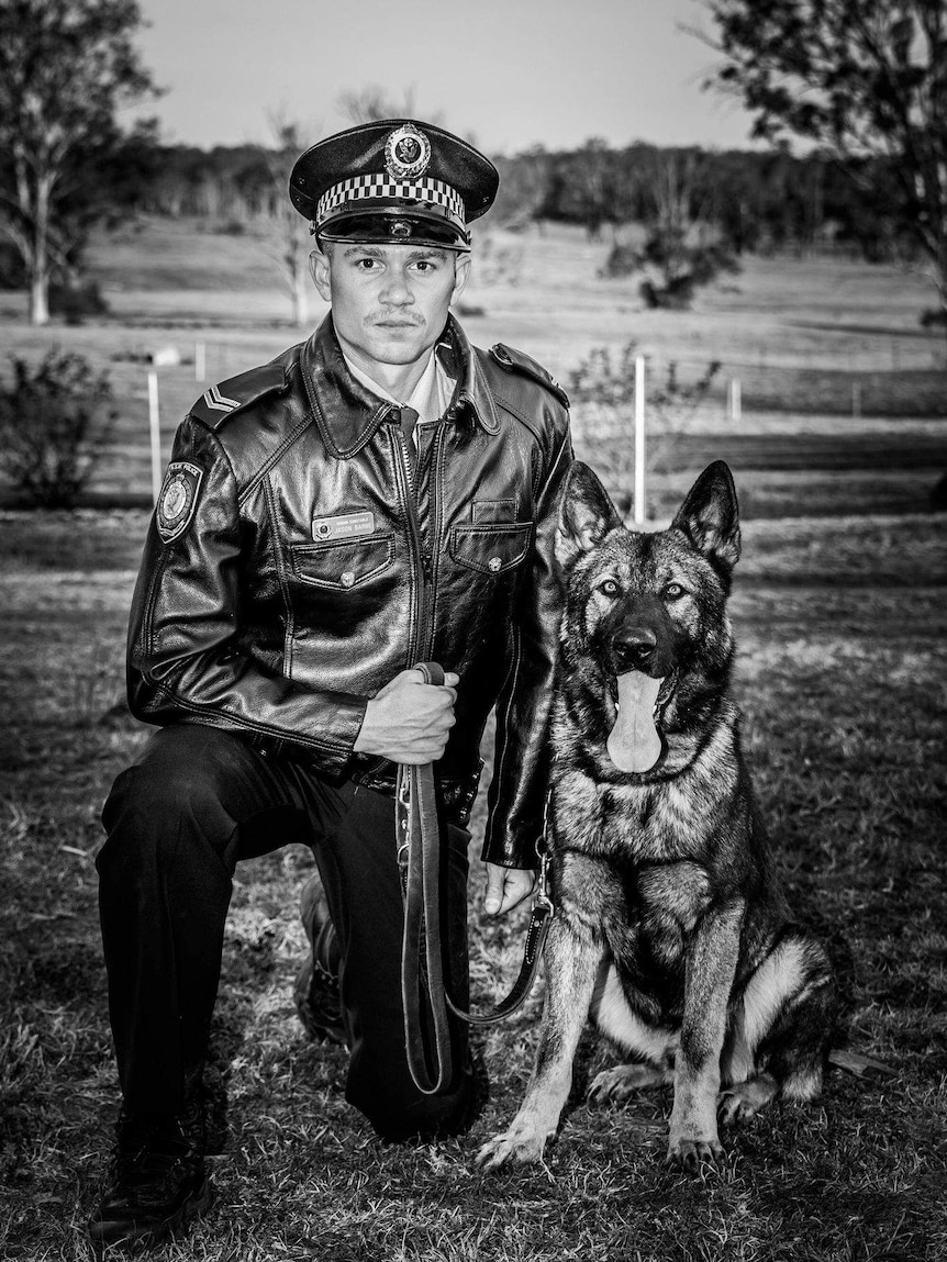 black and white image of man in police uniform with hat kneeling next to large dog with tongue out