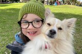 A child wearing a green beanie and glasses hugs a dog as they both look into the camera.
