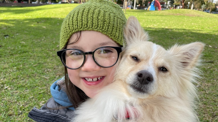 A child wearing a green beanie and glasses hugs a dog as they both look into the camera.