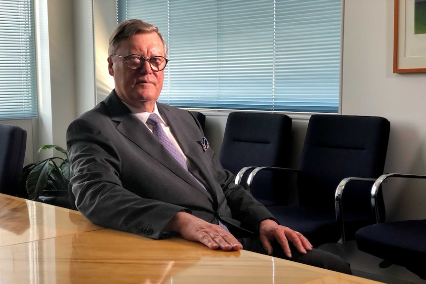 Man in suit and tie reclines in chair in boardroom with hand on wooden table and closed window blinds behind him