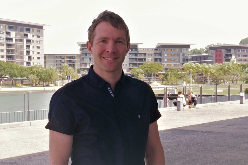 A man with brown hair and a black shirt stands outside. There are buildings and water behind him.