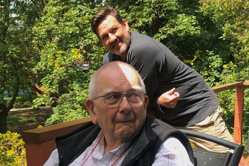 Mike Saclier sits on a chair on an outdoor deck. His son-in-law leans on the bannister of the deck behind him.