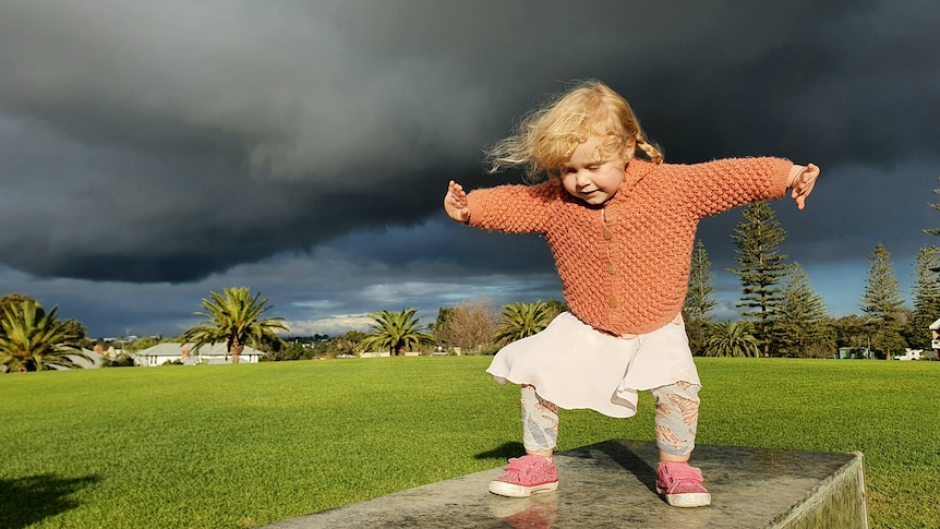 A child in a peach coloured cardigan plays on a stone bench with storm clouds overhead.