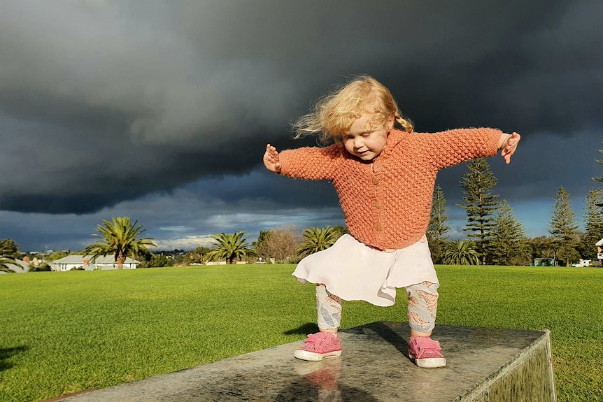 A child in a peach coloured cardigan plays on a stone bench with storm clouds overhead.