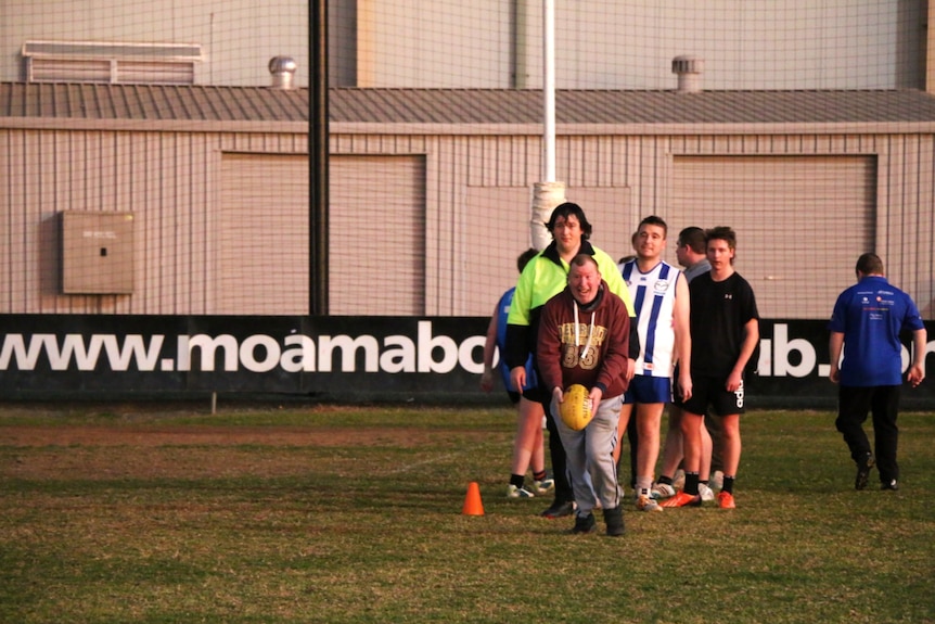 An older man holds a footy, preparing to kick, while other Rockets players line up behind him.