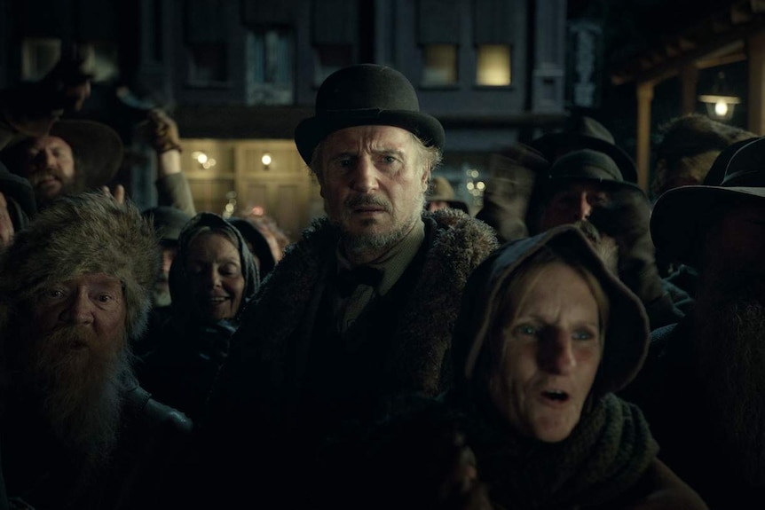 Man in black bowler hat and fur-trimmed coat stands in a crowd of people in Western clothing, looking concerned. Indoors.