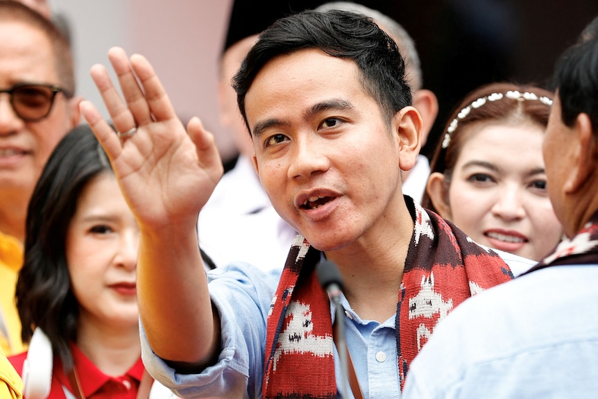 A man wearing a red scarf is surrounded by a crowd of people as he waves to people nearby.