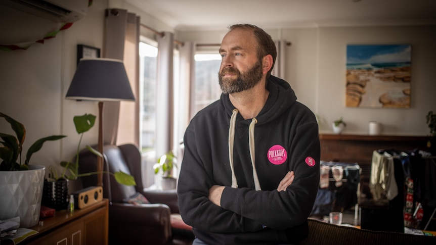  A bearded man stands with crossed arms in an lounge room