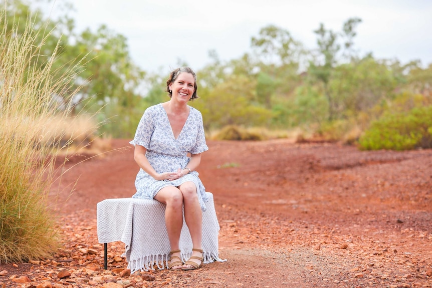 A woman in a floral blue and white dress sits on a bench in the Australian outback.