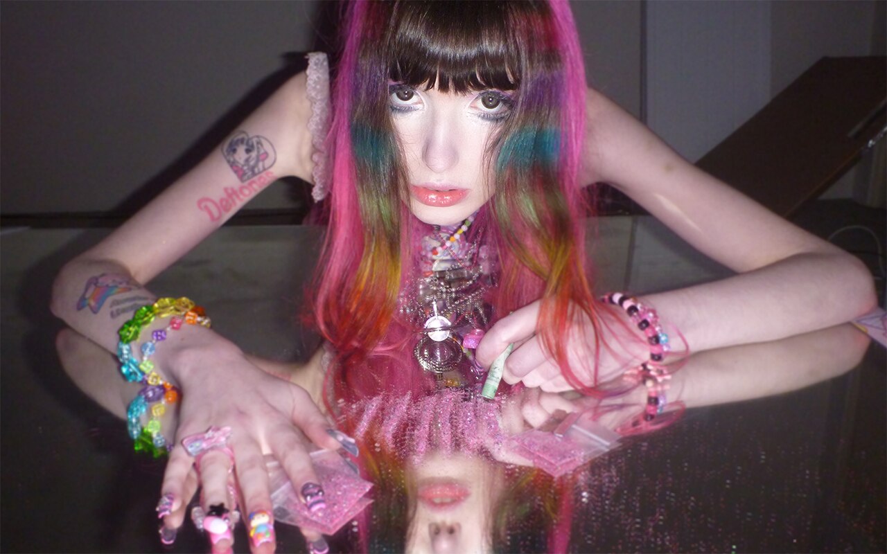 A woman with pink and rainbow hair, big eyes and lots of candy style jewelry lies on a large mirror