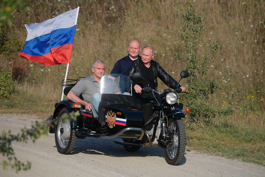 Russian President Vladimir Putin drives a motorbike along a gravel road with a man in the sidecar.