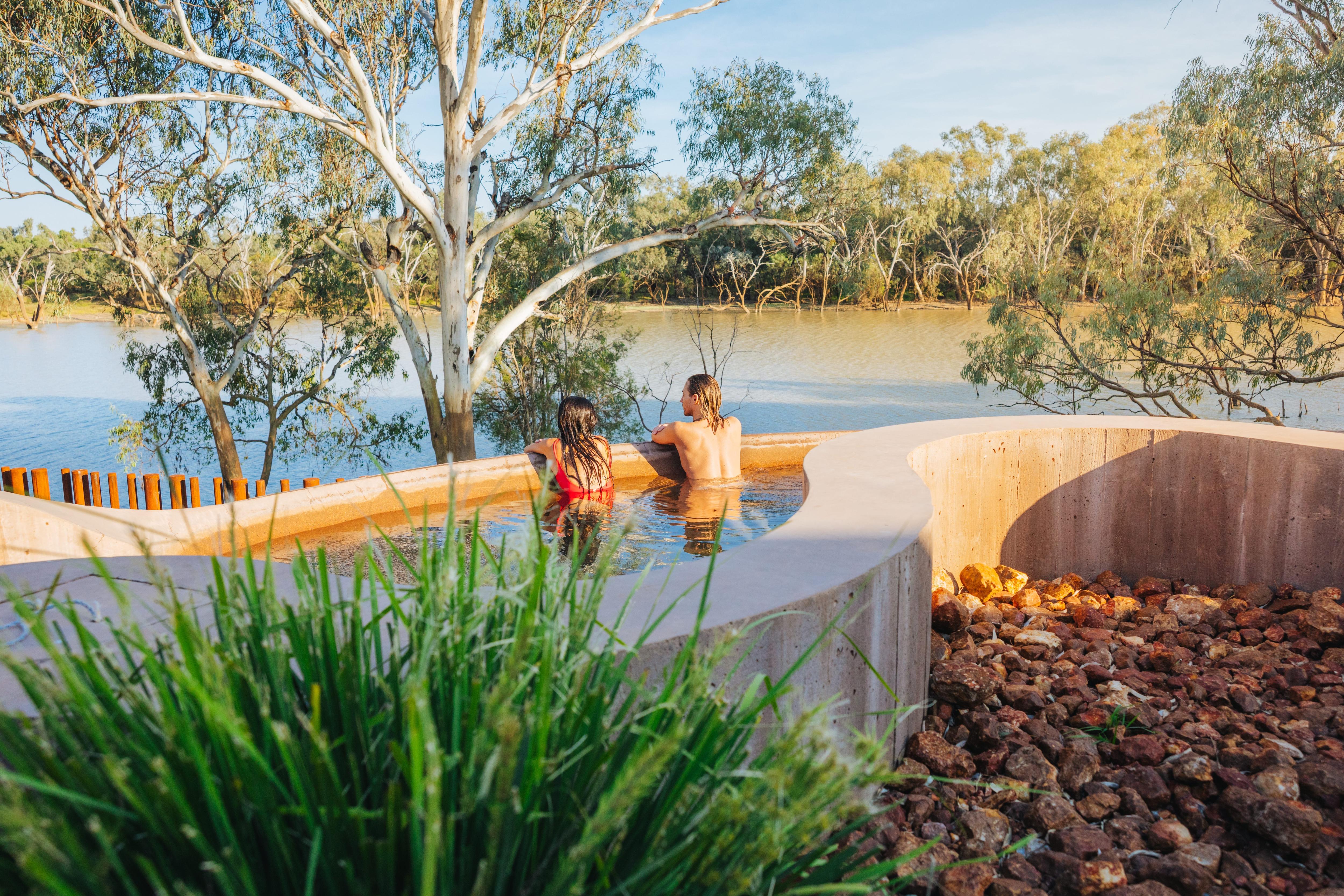 Two people in a hot spring spa looking over a wide brown river with gum trees