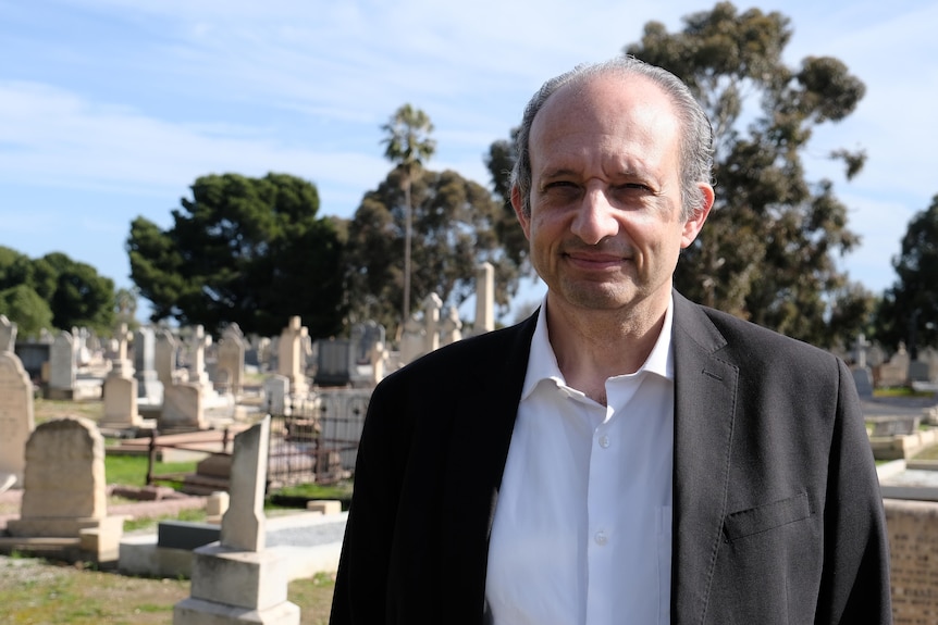 A bald man wearing a suit jacket standing in a cemetery