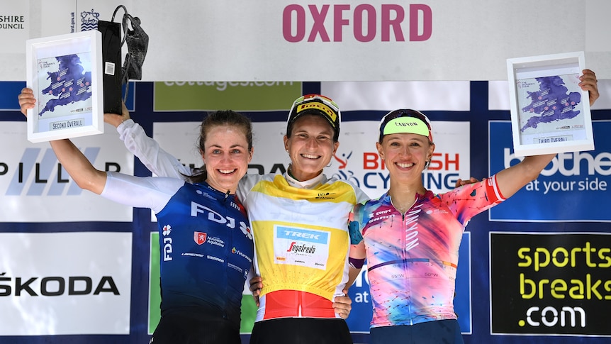 Three female cyclists stand smiling on the podium after a big race, holding their trophies and plaques.