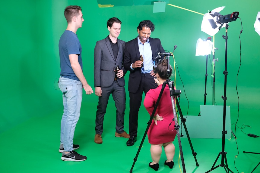 Against a green screen backdrop, one shorter woman is looked down upon by three taller man who laugh and point mockingly.