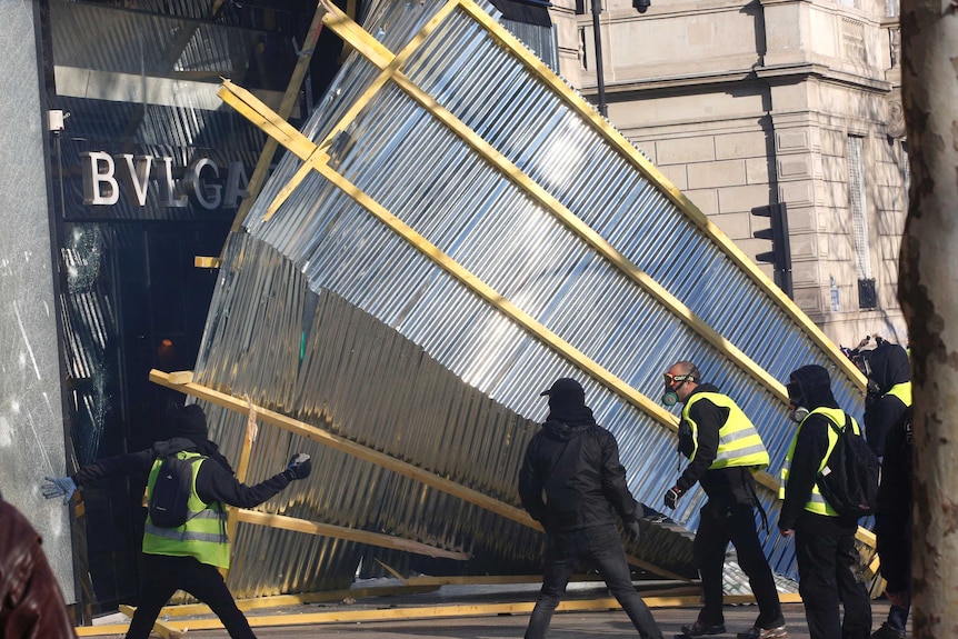 A protestor in a yellow high-visibility vest prepares to throw something near a luxury shop.