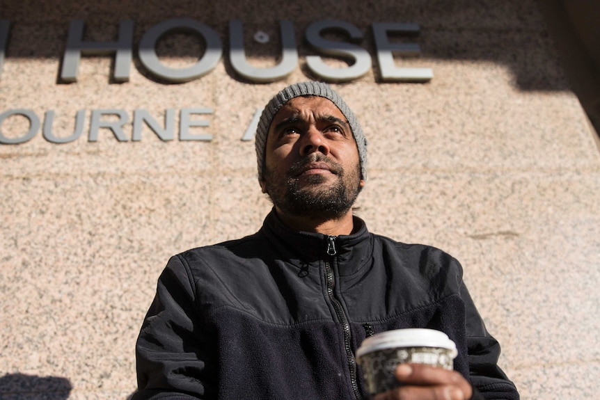 A man holds a coffee up with the word house written on the wall behind him.