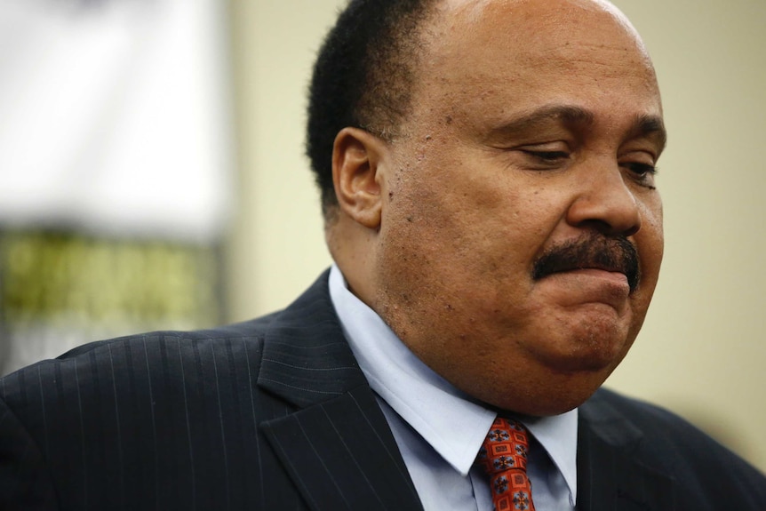 A close up shot of an African American man wearing a suit with a red tie looks down