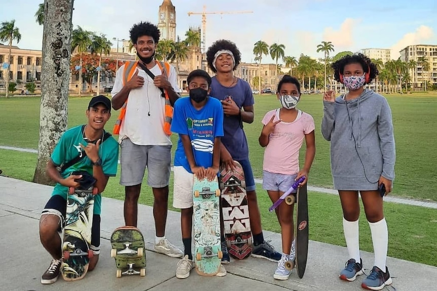 A group of skateboarders stand together in front of the Fijian Parliament House smiling at the camera. 