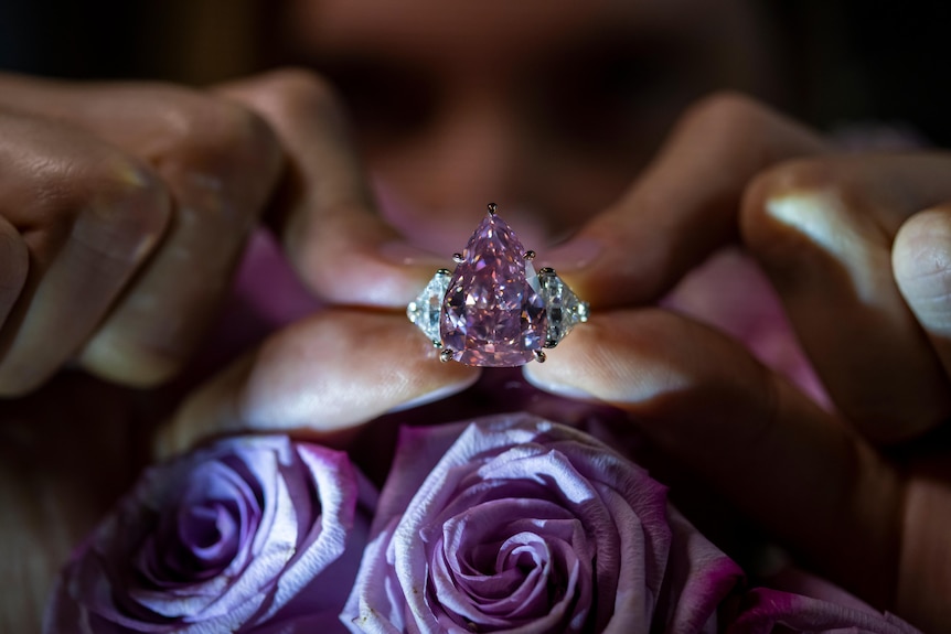Two hands holding a pink pear shaped diamond in front of pink roses