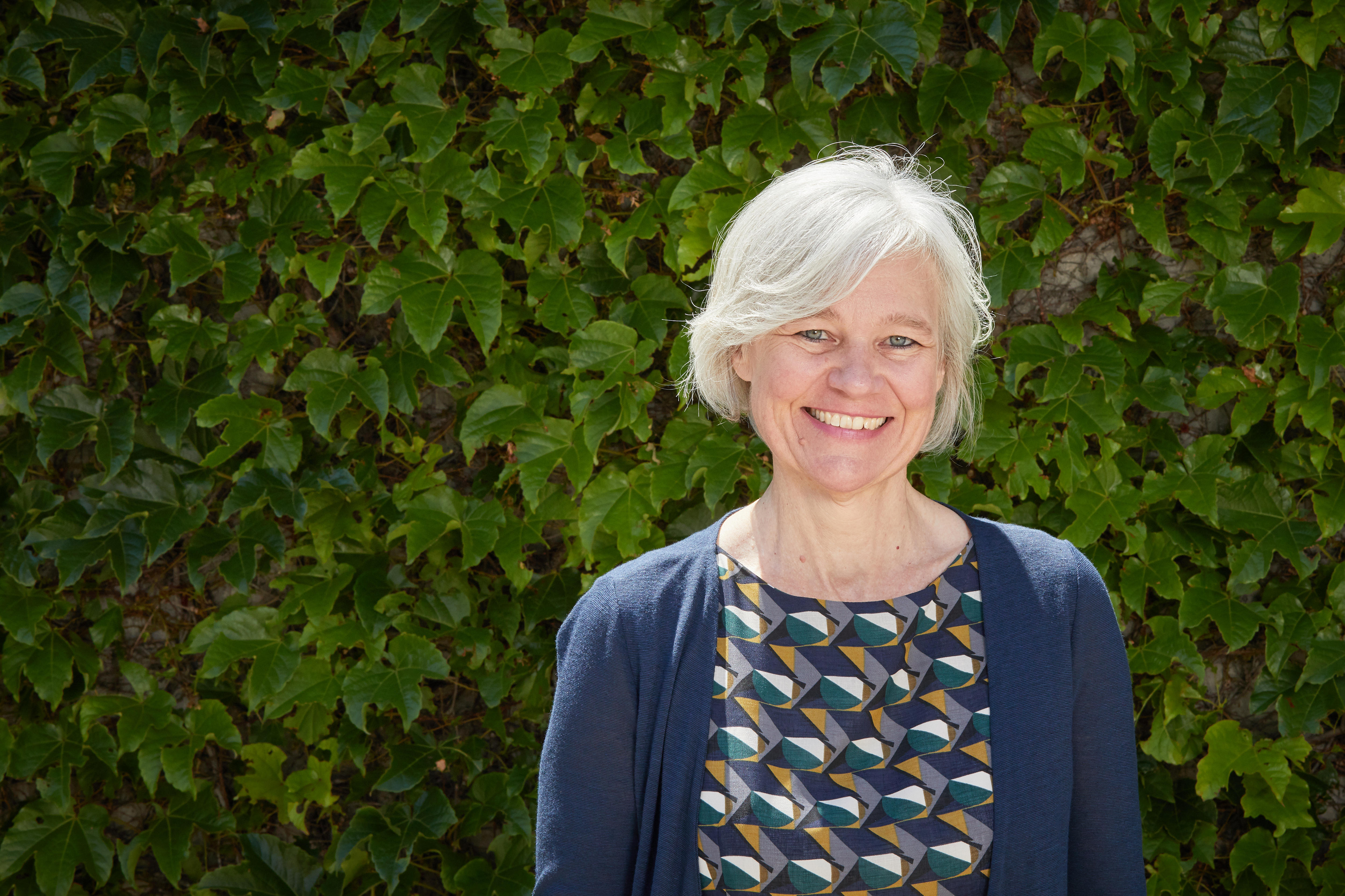 A woman with light white hair and a multicoloured dress and cardigan smiles at the camera.
