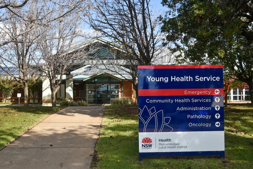 A blue sign reads Young Health Service in front of a tree-lined path to a brick building.
