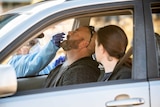A man sits in the front seat of a car next to a woman and his eyes tighten as he receives a nasal swab by a worker outside car.