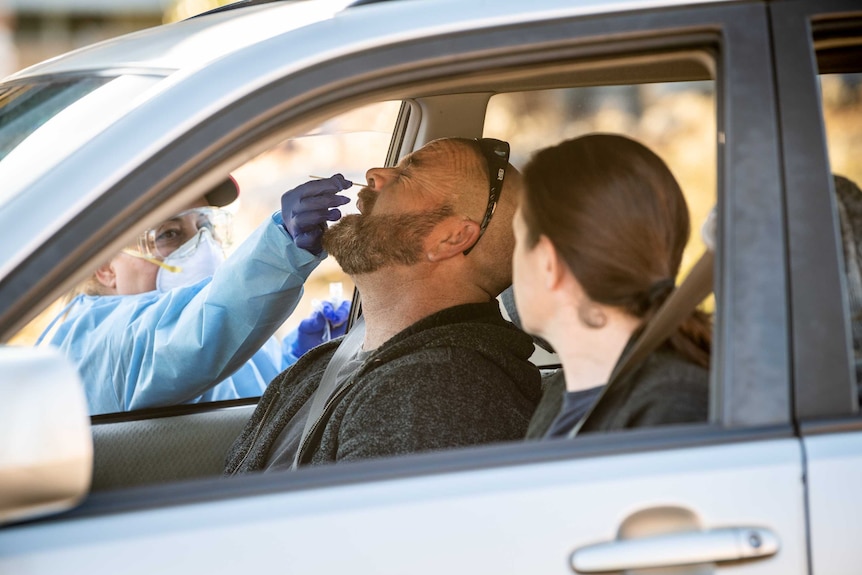 A man sits in the front seat of a car next to a woman and his eyes tighten as he receives a nasal swab by a worker outside car.