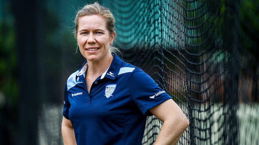 A female cricketer stands next to the nets in training gear staring back at the camera.