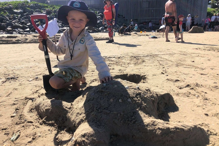 A young boy smiles as he sits next to a turtle sandcastle