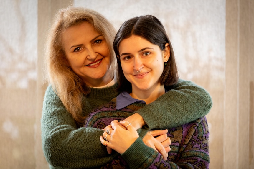 A woman with blonde medium-length hair smiles and wraps her arms around her daughter.