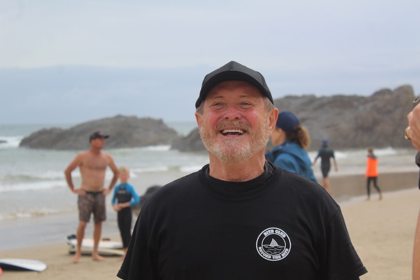 A man stands on a beach, smiling and wearing a cap.