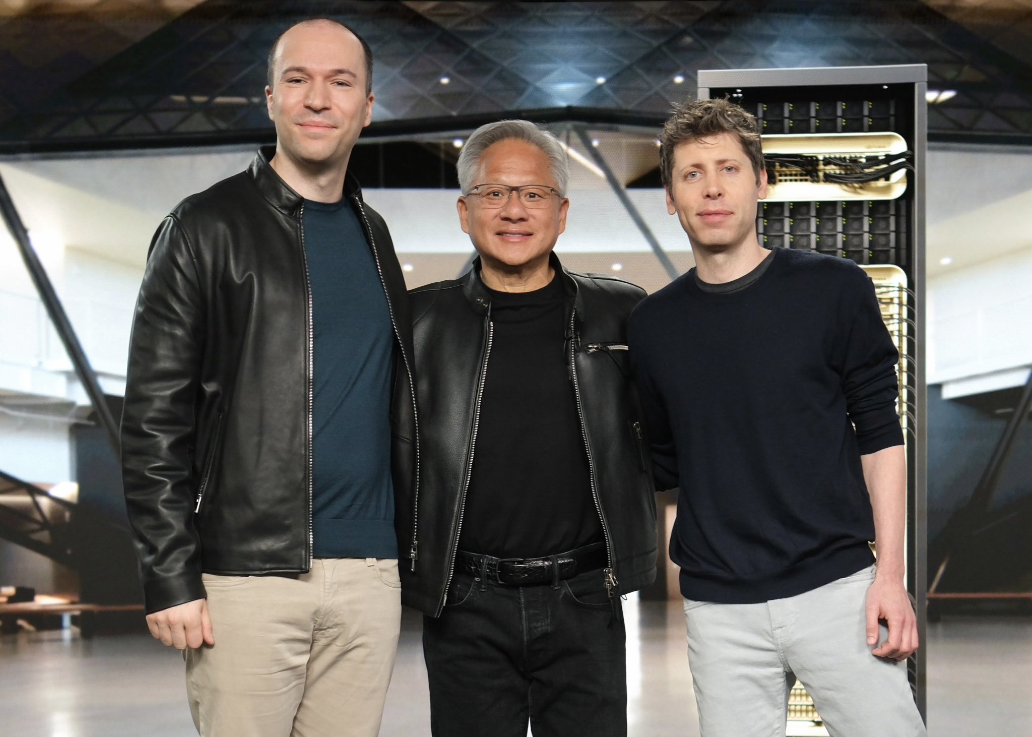 Greg Brockman, Jensen Huang and Sam Altman stand shoulder to shoulder and smile for a group photo.