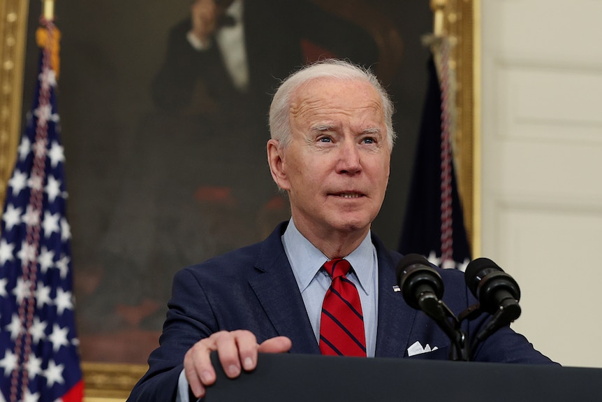 An elderly man in a dark suit stands a lecturn in front of a painting and makes a speech